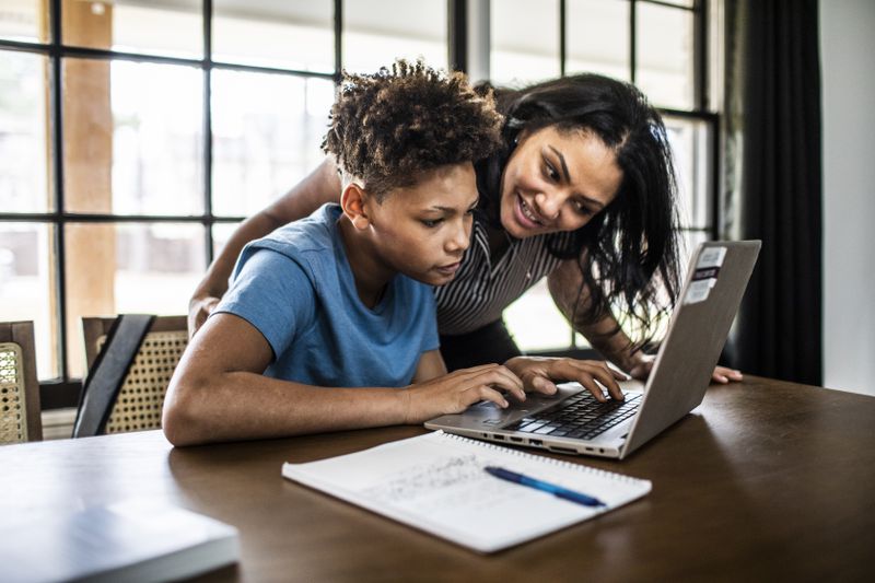 Adult woman helping younger man on laptop at a dining room table.