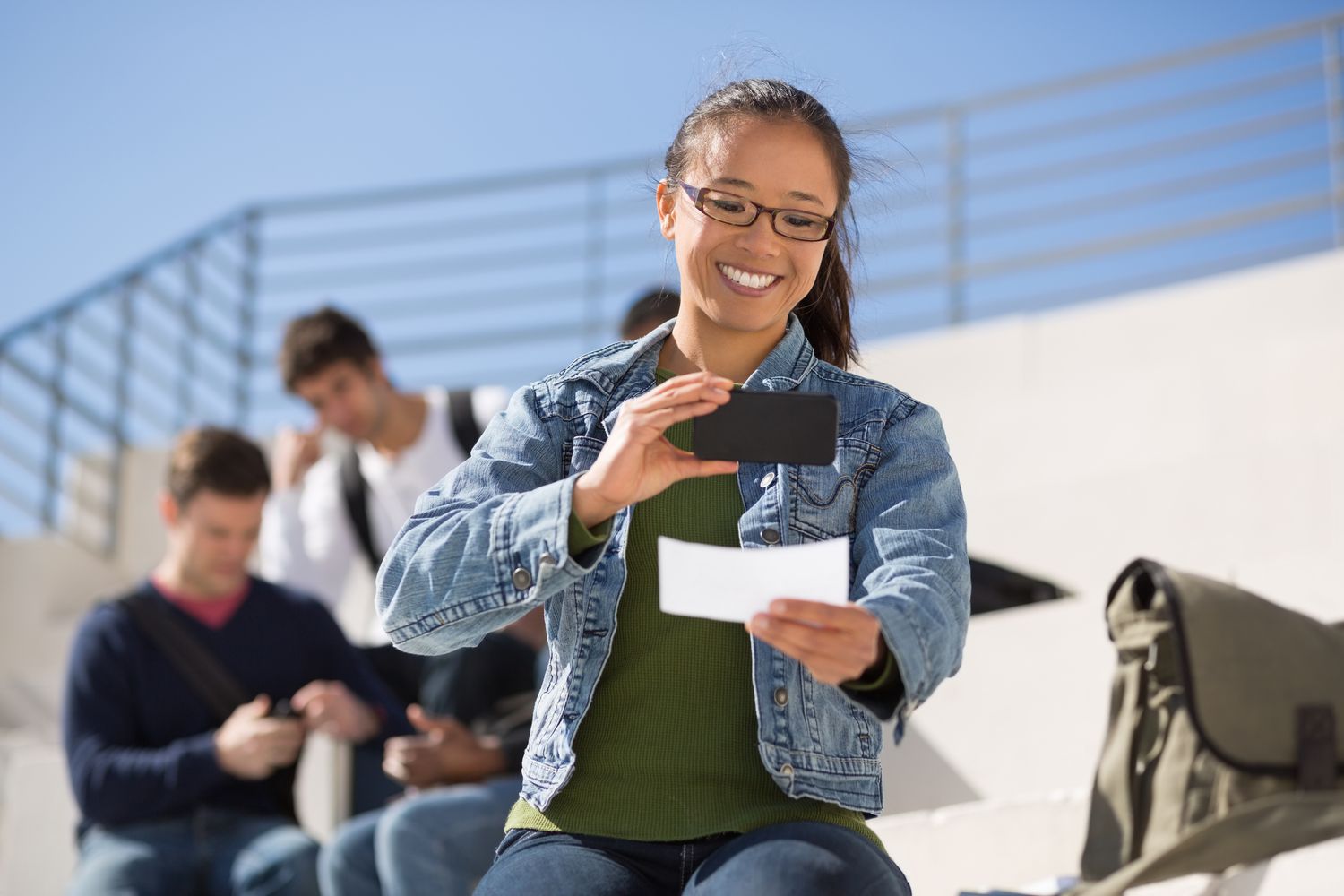 A young woman deposits a check with a mobile phone while sitting outside on bleachers.