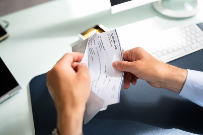 A worker pulls their paycheck out out of an envelope while sitting at their office desk.