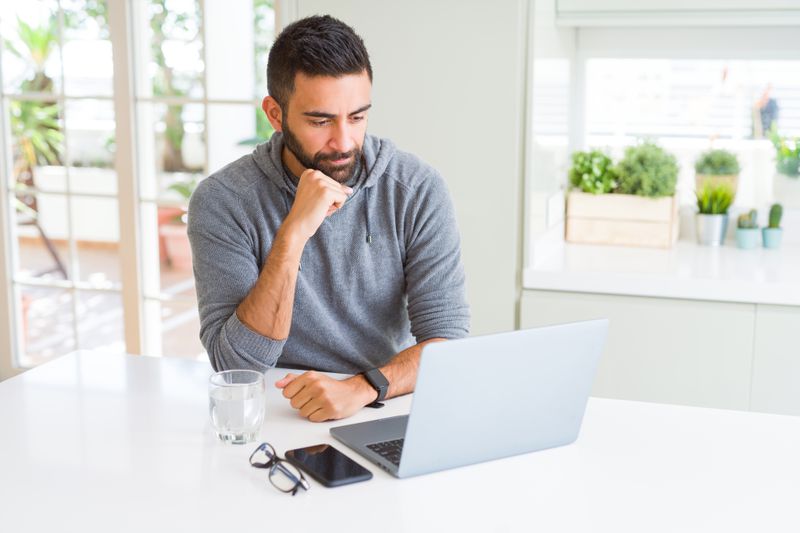 Man in his 30s sitting at his kitchen table and pondering something on his laptop
