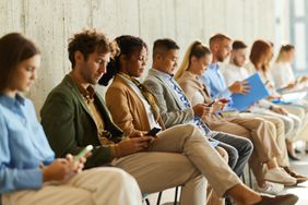 A group of people seated in a row in a hallway, some looking at their phones and others holding documents