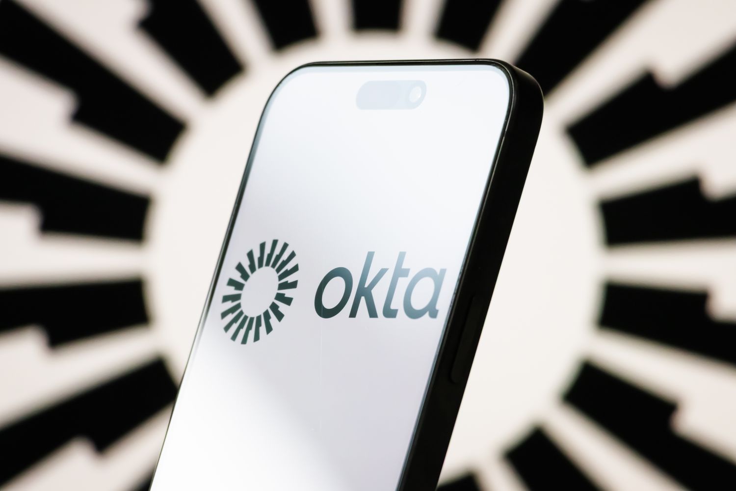 The logo of Okta is displayed on a smartphone screen, with the company's stylized black-and-white branding visible in the background