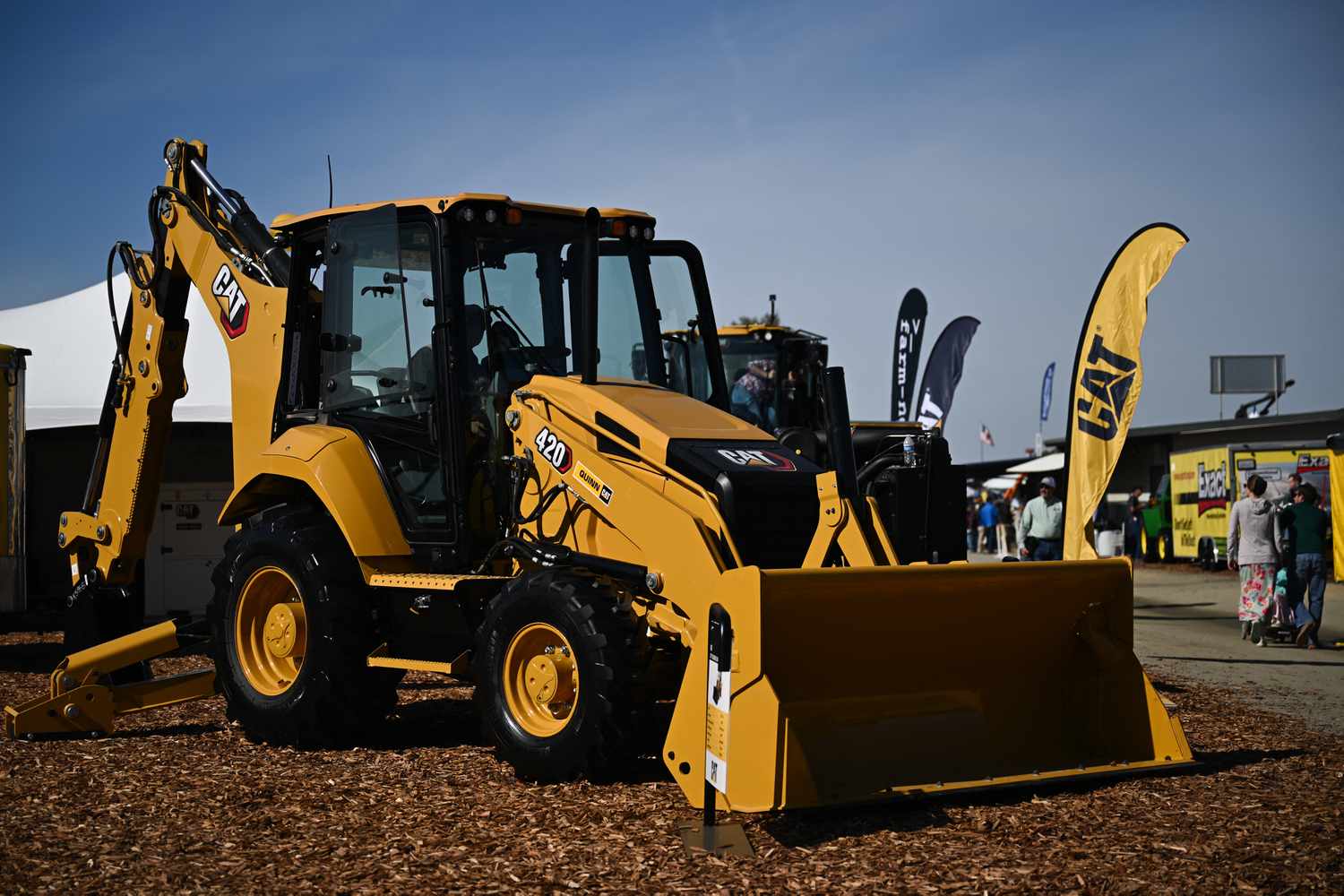 A yellow Caterpillar backhoe sits on a patch of mulch, with a banner bearing the company's black and yellow CAT logo to the right.