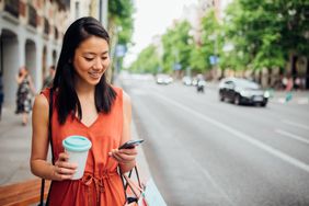 A woman in an orange dress stands on a street with a coffee and smartphone in hand