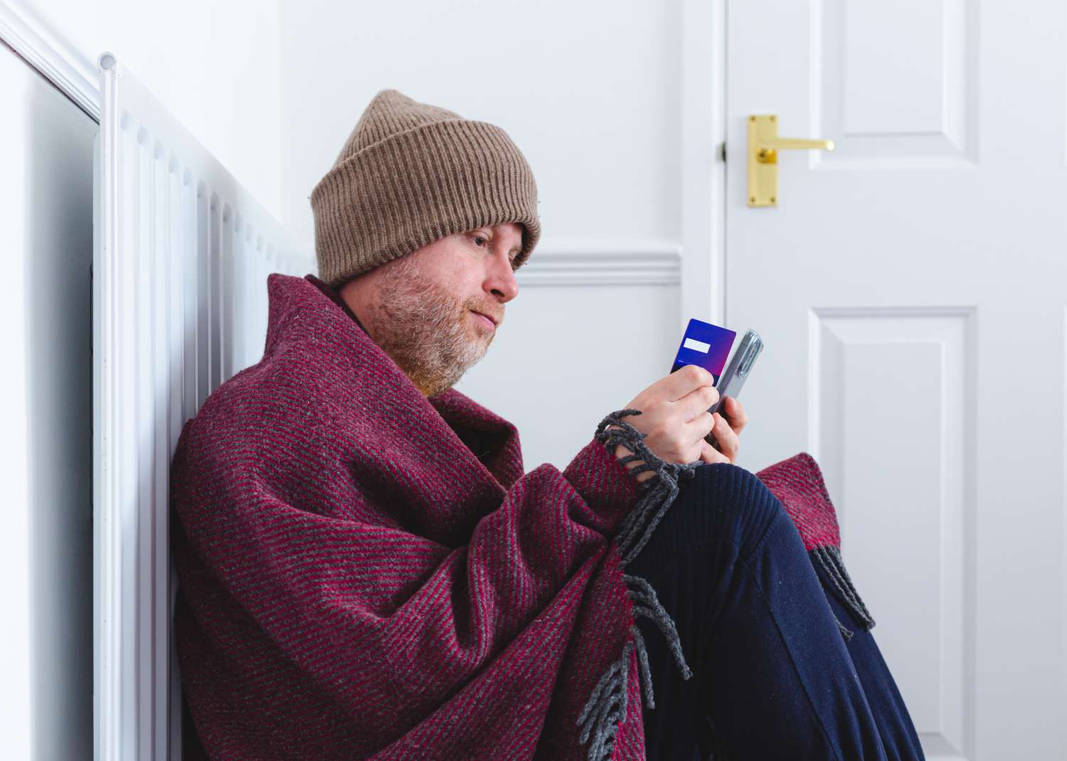 A person wrapped up in layers of clothing sitting on the floor and considering freezing the credit card in hand.