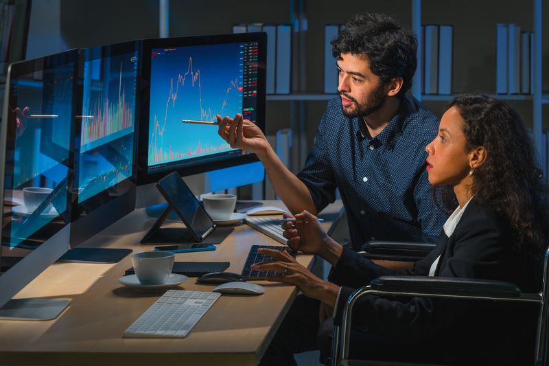 Two stock traders sit in a darken office analyzing price charts displayed on monitors to decide on pricing for future contracts.