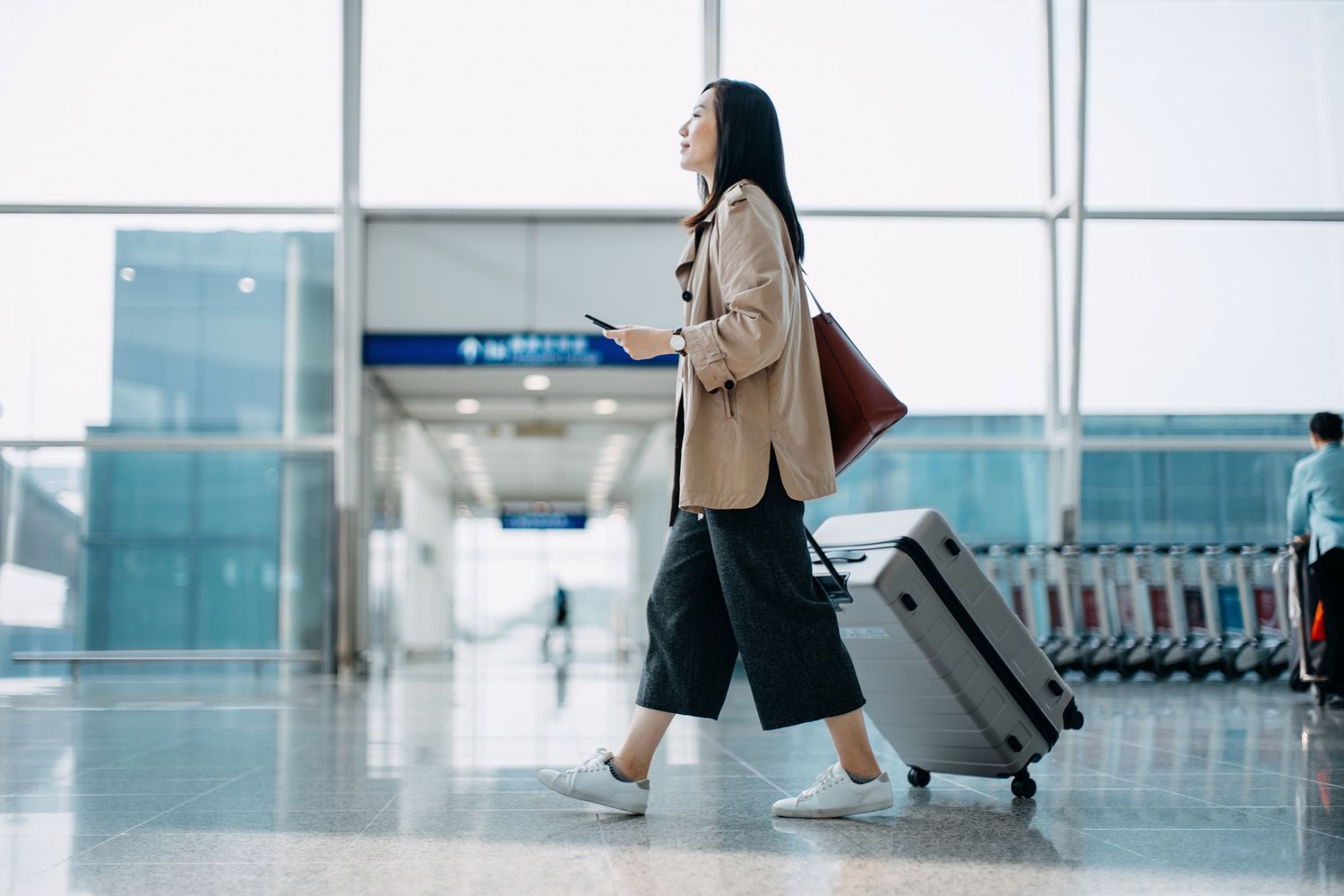 woman carrying suitcase and holding smartphone on hand, walking in airport terminal