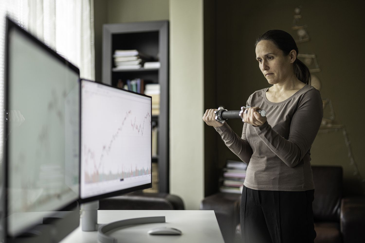 Female trader working at home, standing in front of two monitors and exercising.