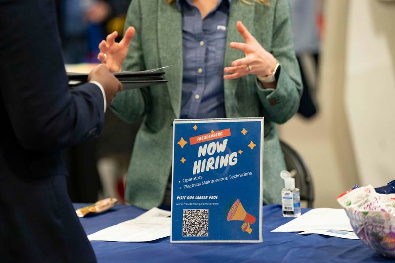 Person speaking to a job recruiter at a hiring event, with a "now hiring" sign on the table