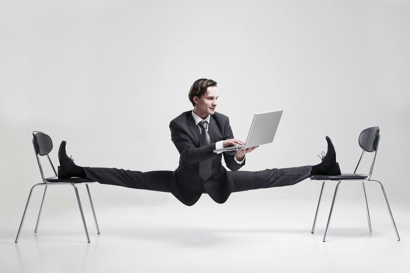 Young businessman, wearing dark grey suit, doing the splits balanced on two black chairs whilst holding a laptop.
