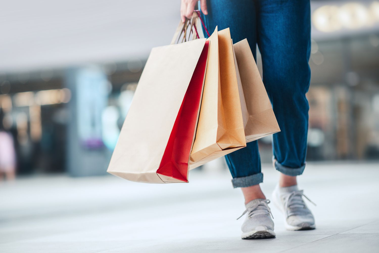 Bottom cropped legs of a person wearing jeans and sneakers and holding three paper shopping bags
