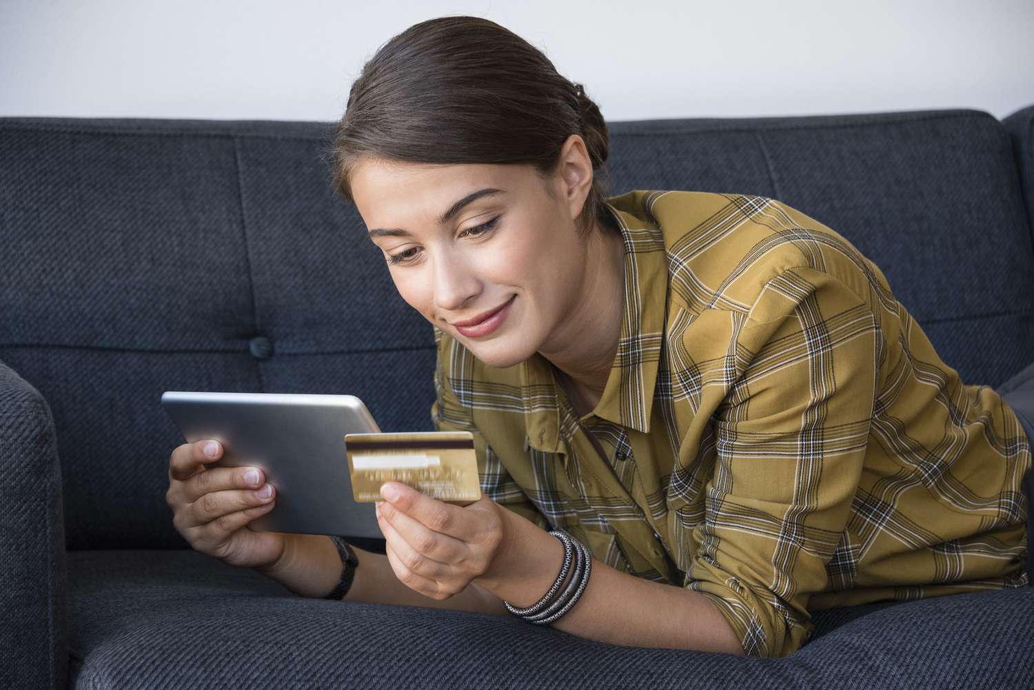 A smiling woman lays on a couch, looking at a bank card and a tablet.
