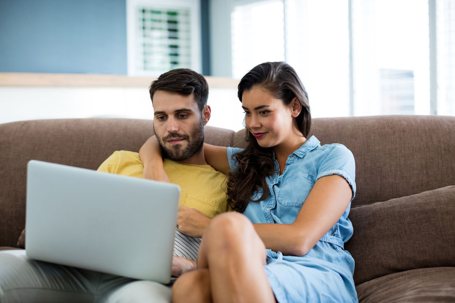 A man in a yellow shirt and a woman in a blue dress sit on a couch looking at a laptop