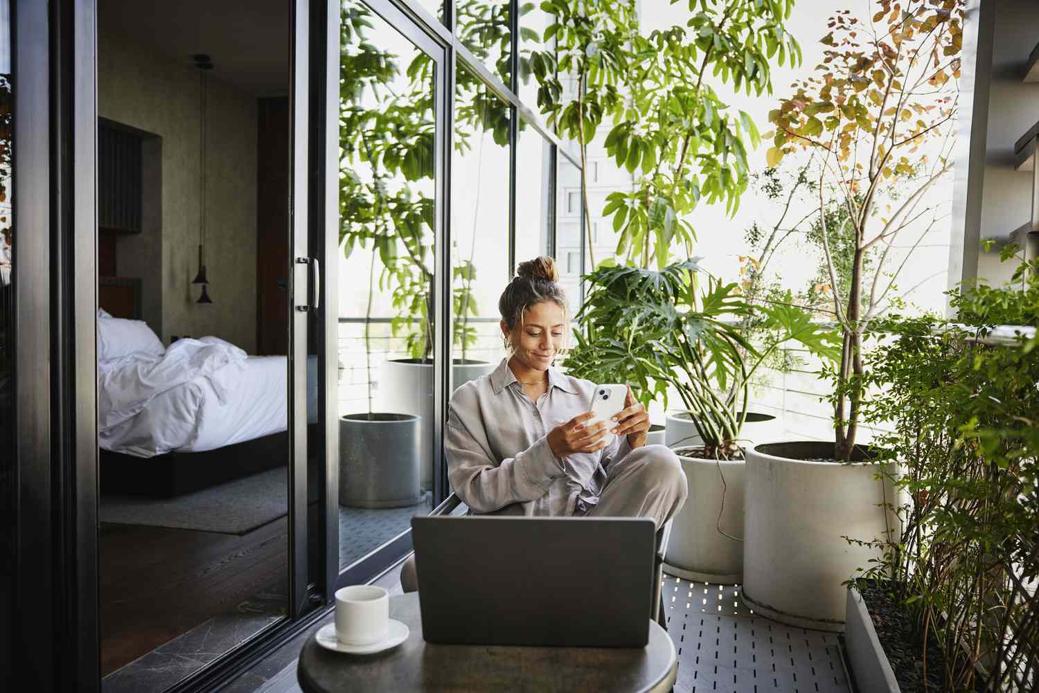 Person in pajamas seated on a balcony using a phone, surrounded by plants, and a laptop on a small table