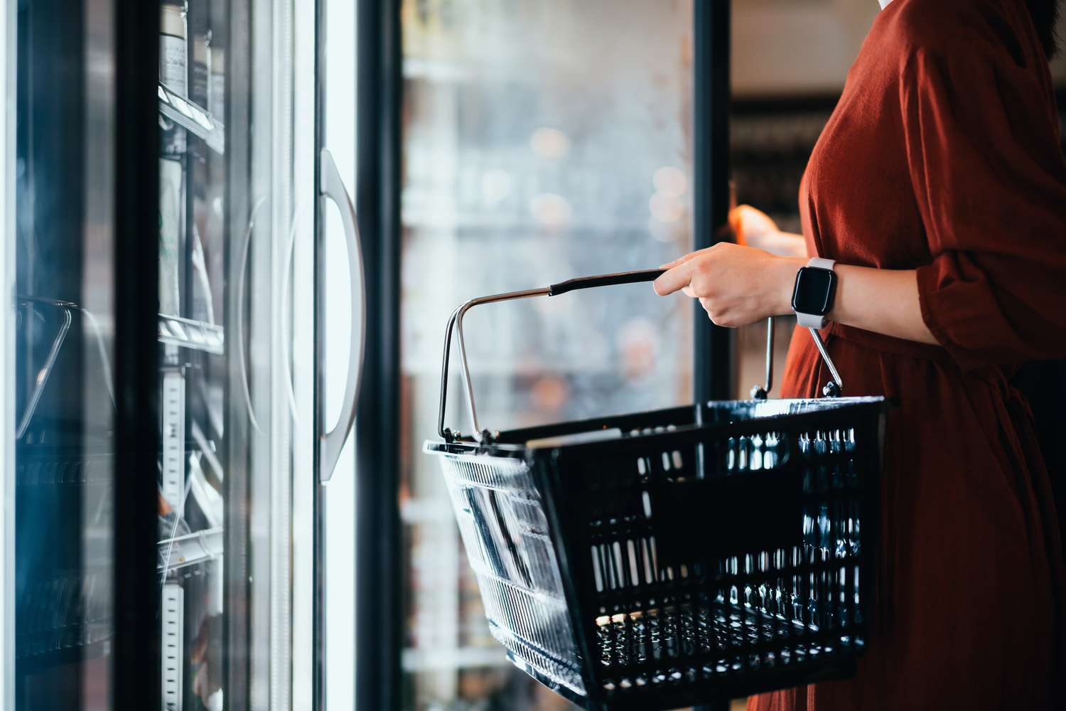 A person shops for food with a basket in a store's refrigerated aisle. 
