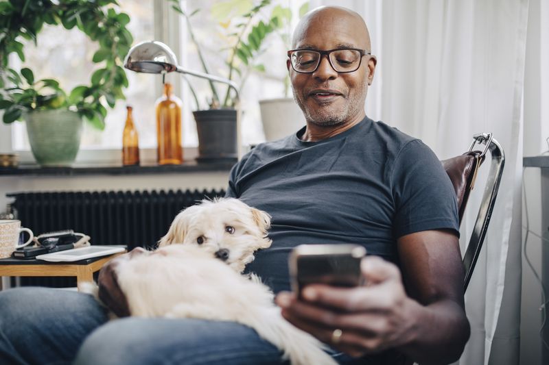 Smiling retired senior male using smart phone while sitting with dog in room at home.