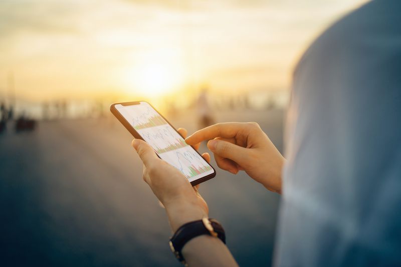 Close up of woman's hand checking financial stock data on a mobile device by the pier at golden sunset