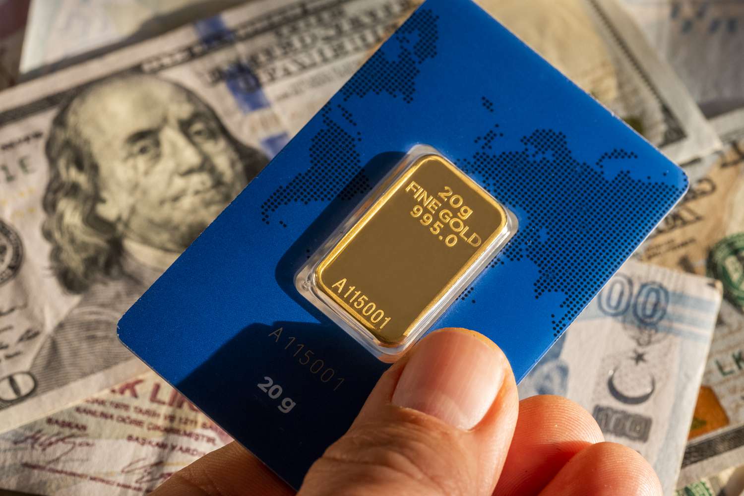 A hand holding a small gold bar with various international currency bills 