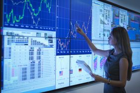 Woman standing in front of a wall of computer monitors, pointing to a chart. 