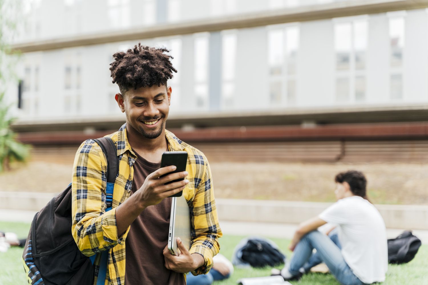 College student on college campus walking and looking at smart phone holding laptop.