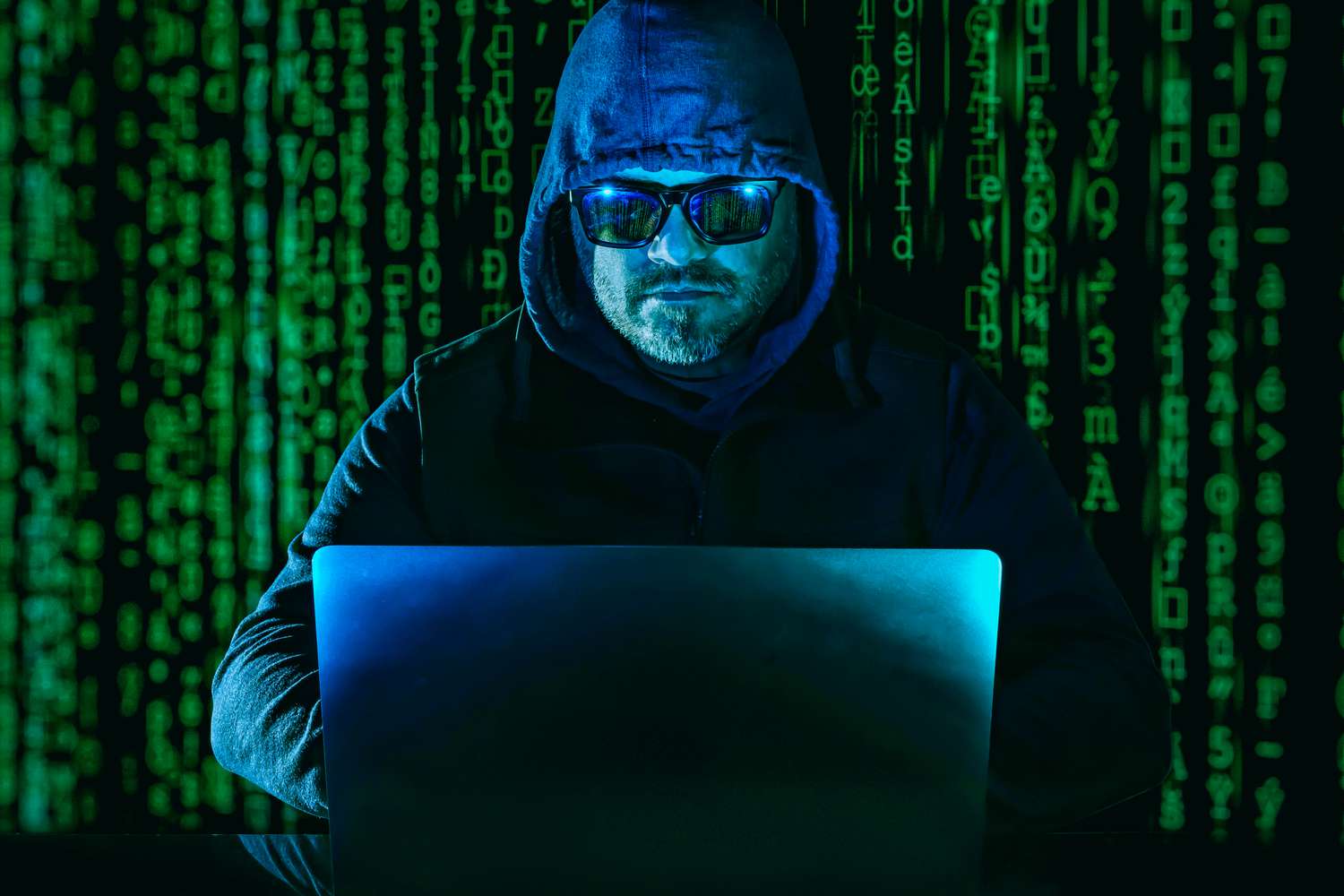 An anonymous person wearing a hooded jacket and sunglasses uses a laptop in front of scrolling rows of cryptocurrency symbols