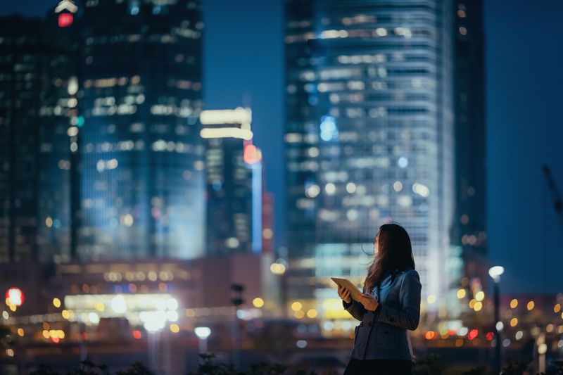 Young businesswoman using digital tablet looking back over at illuminated skyline