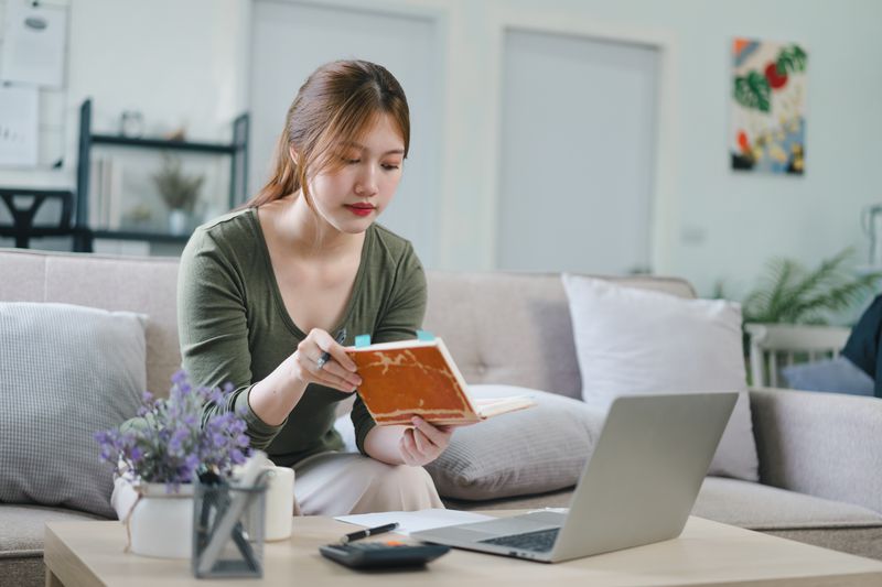 Person sits on a couch with a laptop on a table before her. She is holding a notebook.