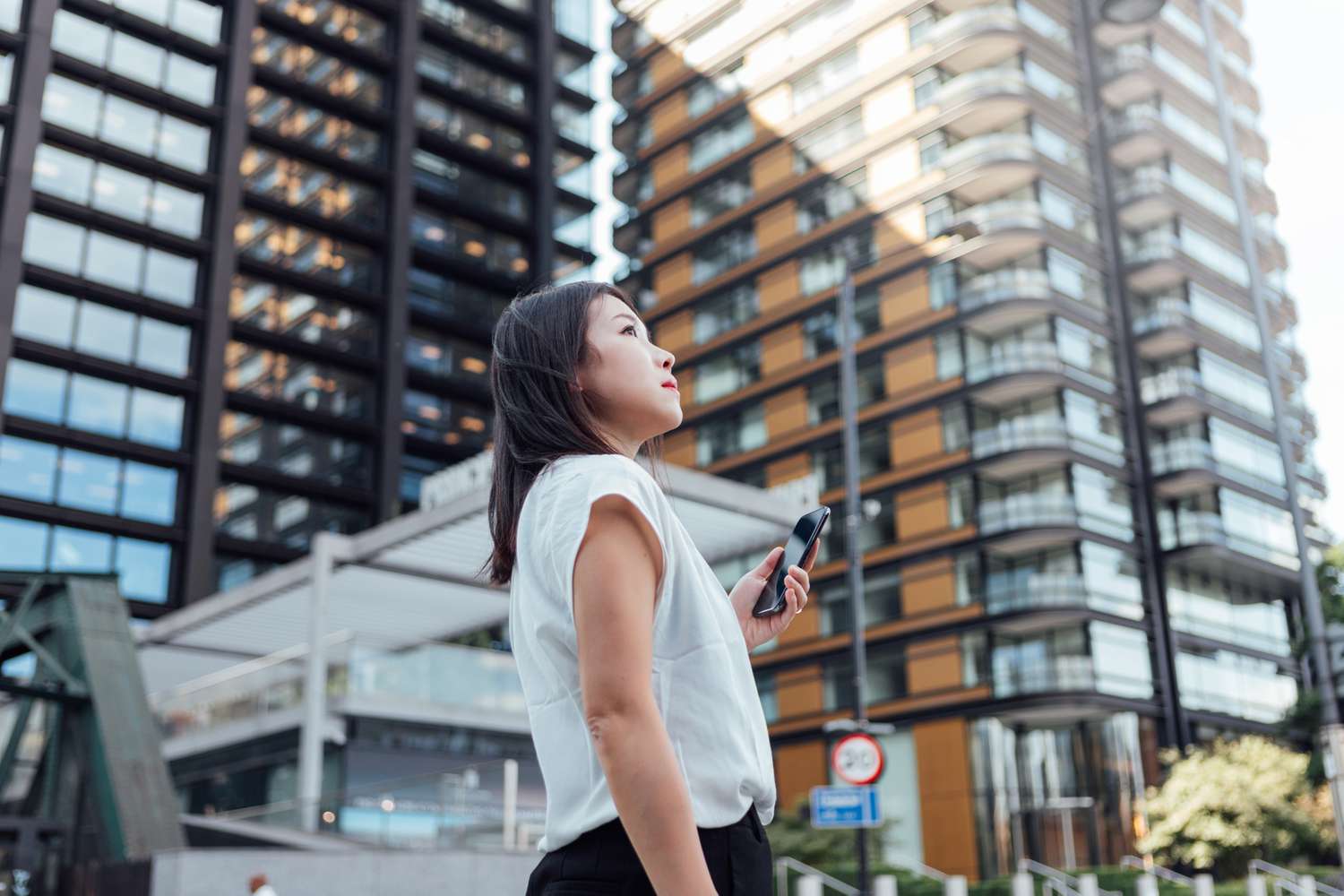 Young woman with a smartphone stands in front of a skyrise building. 
