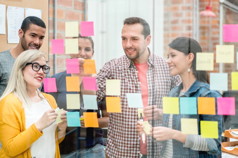 A group of coworkers in an office place sticky notes displaying customer demographics and personality into groups on a large glass room divider.