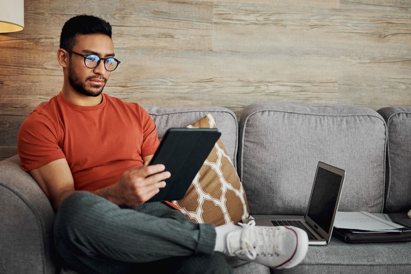 A person sitting on a couch holding a tablet, with a laptop and notebook nearby