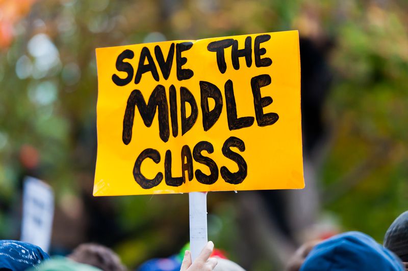 A hand holds a poster sign that reads "SAVE THE MIDDLE CLASS" in black marker on yellow paper in the middle of a crowd.