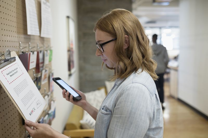 Female freelancer with smart phone checking message board in office.