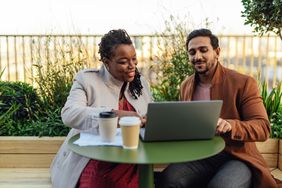 Two people discussing finances while looking at a laptop. They are sitting on a patio and drinking coffee.