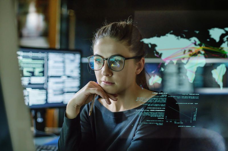 Stock image of a young person, wearing glasses, surrounded by computer monitors in a dark office. In front of her there is a see-through displaying showing a map of the world with some data.