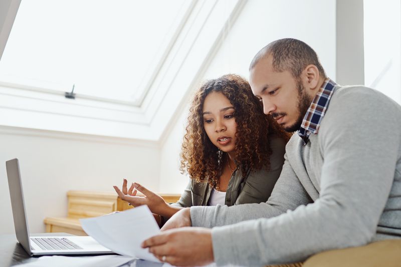 Two people look at paper documents and a laptop at home.