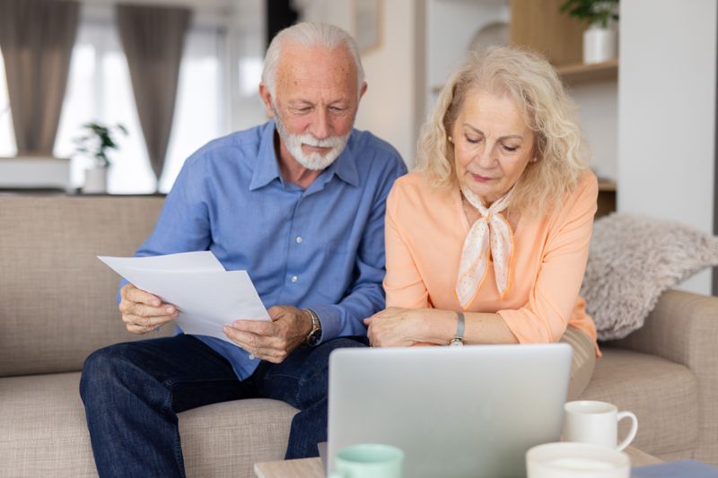 Older couple sitting on their sofa and looking together at a laptop and financial documents