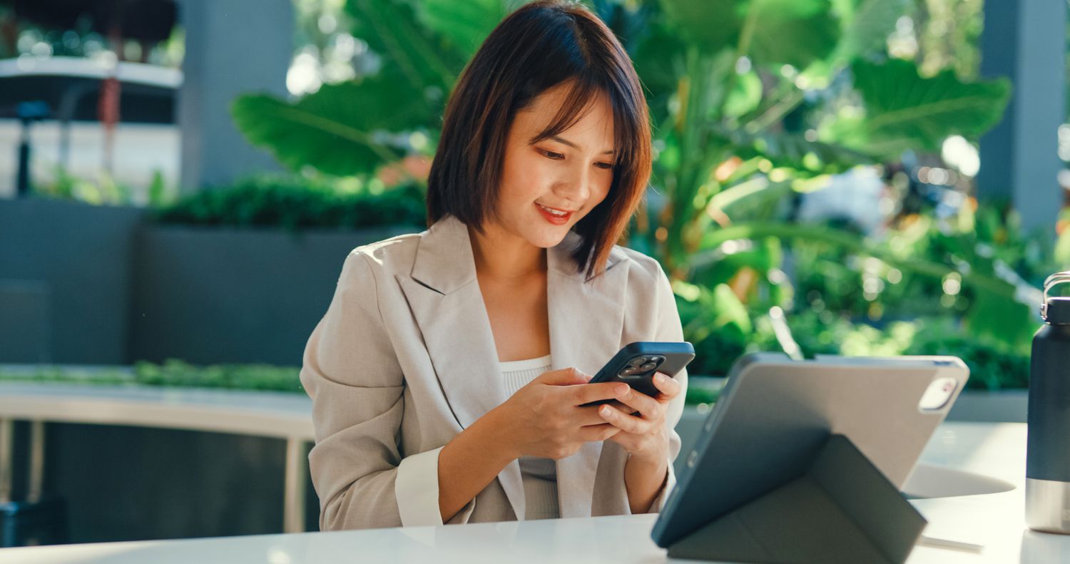 Woman looking at her mobile phone with a laptop on the desk in front of her.