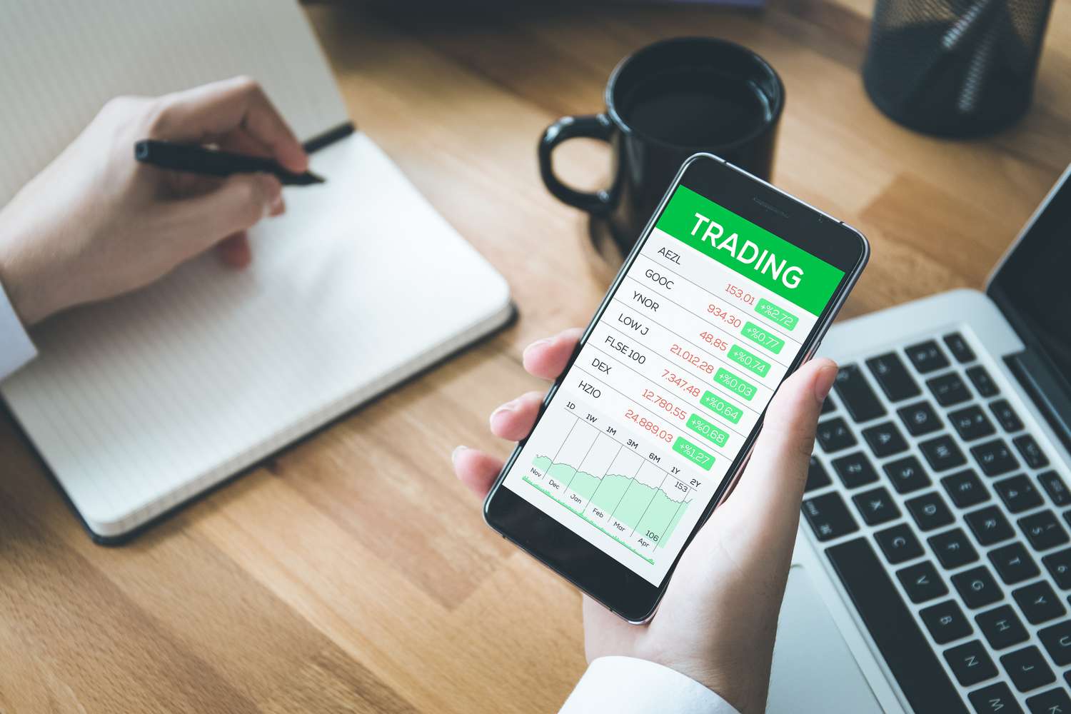 An investor holds a cellphone displaying a list of stocks they are following in one hand while writing notes in a notebook with their other hand. 