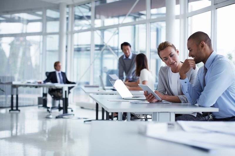 Colleagues discussing over a digital tablet in a large, white office space with co-workers in the background
