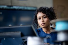 A woman sits before a laptop while a fuzzy monitor sits in the background. 