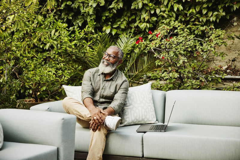 Medium wide shot portrait of smiling senior man with laptop sitting in garden on rooftop deck