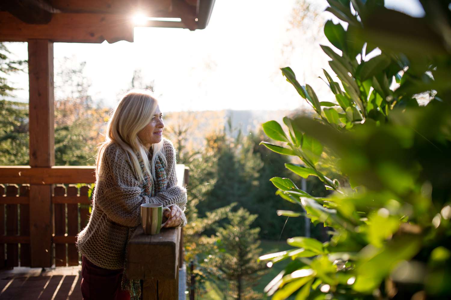 A mature woman holding a coffee mug in sunlight while standing on a dek overlooking a wooded area. 