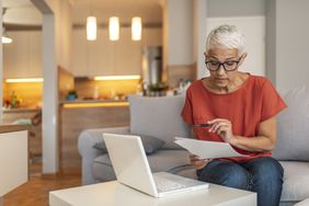 A woman in an orange shirt and jeans sits on her couch looking at a document with a laptop open in front of her on the coffee table.