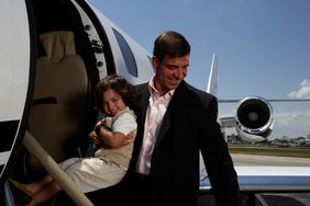 A man holding a child while standing near the doorway of a private jet, seen outdoors with part of the aircraft and sky in the background