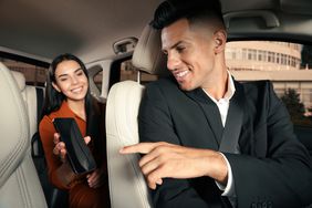 A man in a car suit pointing at a smiling woman holding a smartphone in the backseat