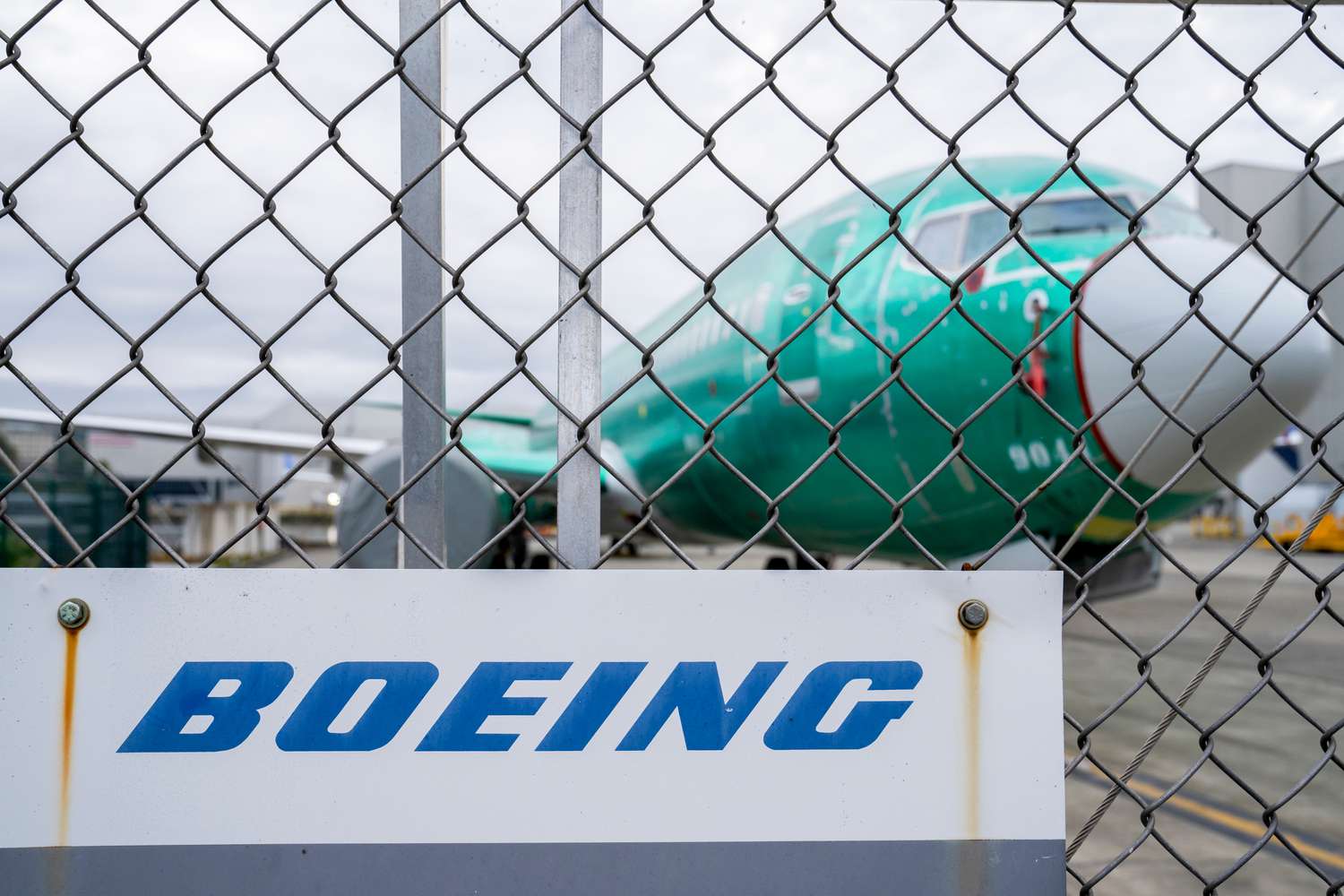 The Boeing logo is seen on a sign in front of a green plane that is under construction.