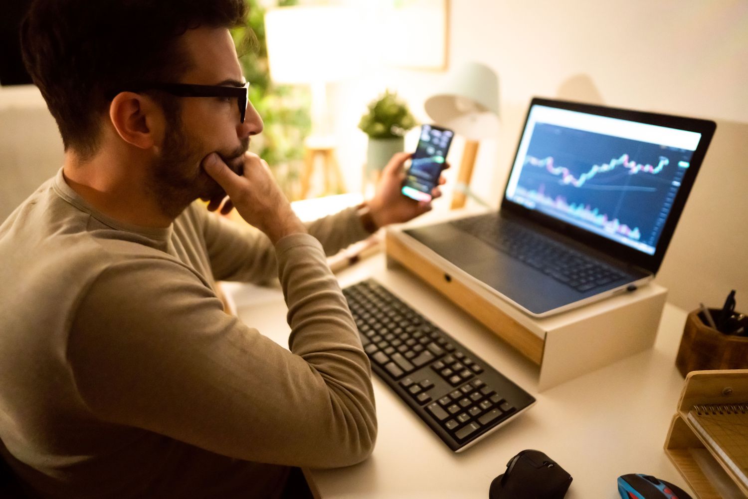 Man looking at market data on a smartphone and a laptop screen