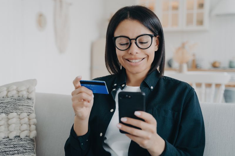 A smiling woman sits on a couch, holding a smartphone and a payment card.