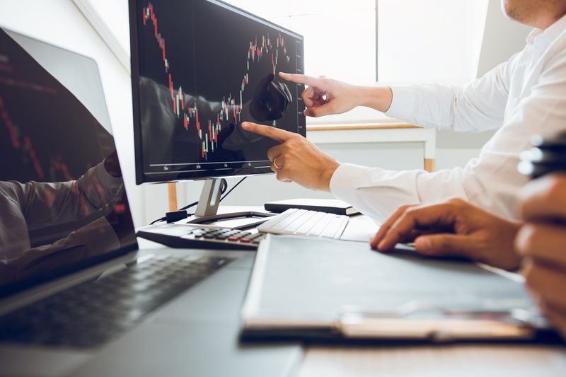 A person wearing a collared shirt points at a stock chart on a computer screen, while a person in the foreground sits in front of a laptop.