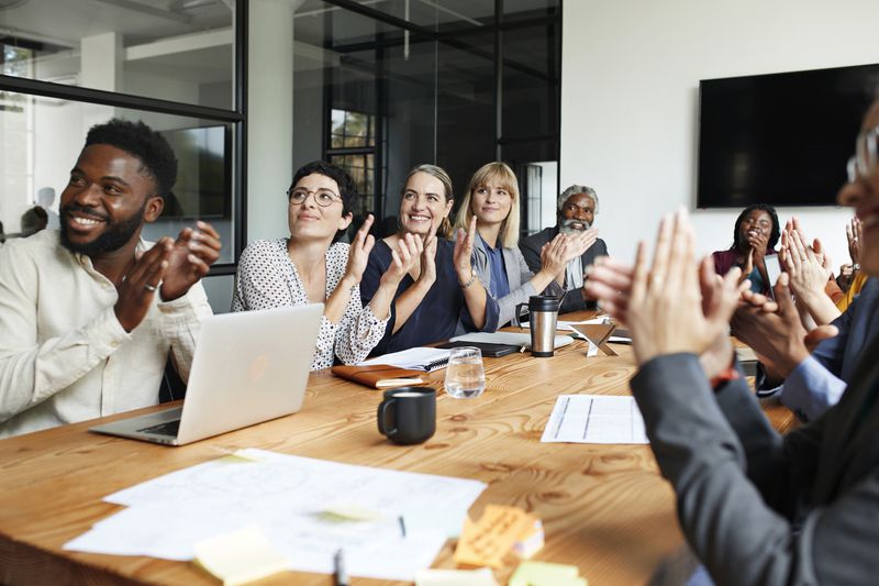 Smiling male and female business people clapping while sitting by conference table in board meeting